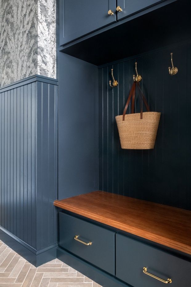 A compact, richly detailed entryway with deep navy-blue built-in cabinetry and vertical beadboard wall panelling. A warm timber bench seat tops lower drawers, with brass handles and wall hooks holding a woven basket. The upper wall includes patterned botanical wallpaper framed by blue panelling, and the floor is finished in light neutral herringbone tiles.