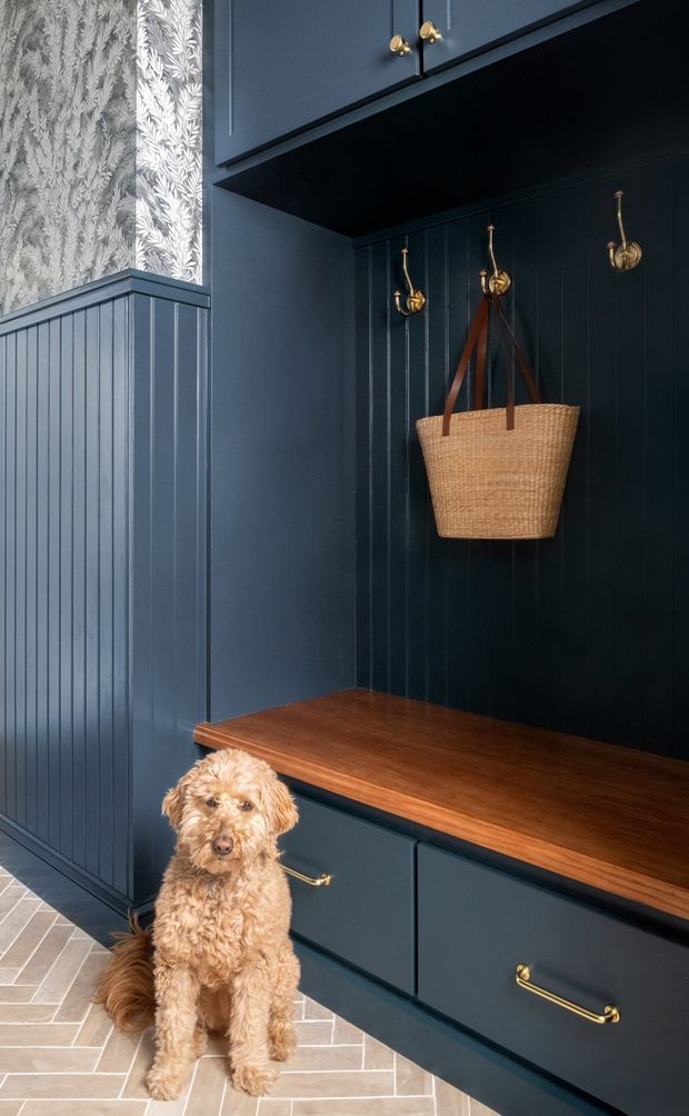 A dark blue built-in entryway or mudroom with vertical beadboard wall panelling, upper and lower cabinetry, a warm timber bench seat, and brass hooks and drawer pulls. A woven basket hangs on the hooks, while patterned botanical wallpaper appears above the wainscoting. The floor is laid with light herringbone tiles, and a dog sits in front of the joinery.