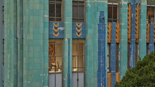 Close-up exterior view of a distinctive building facade clad in turquoise and blue glazed tiles, with vertical fluted surface treatment, narrow windows, and geometric chevron and lattice-like decorative detailing in blue, ochre, and timber tones. Dense greenery partially obscures the lower right edge.