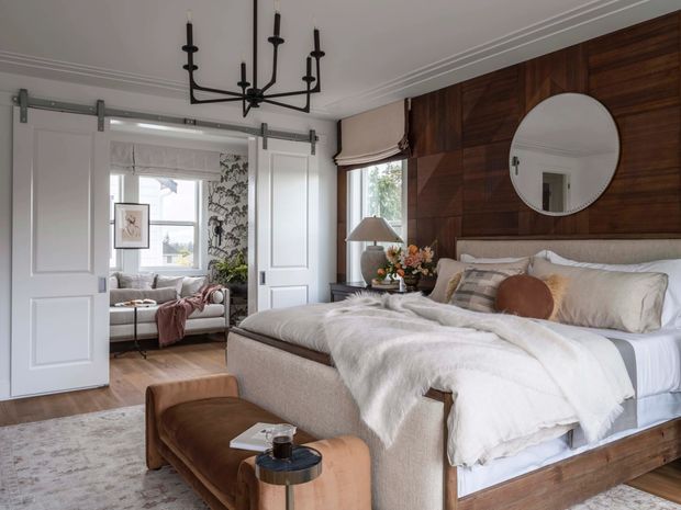 A refined bedroom with a large upholstered bed, layered white and neutral bedding, and a dark timber feature wall behind the bed. White sliding barn-style doors open to a bright window nook, while a black linear chandelier, round wall mirror, bedside lamp, upholstered bench, and small side table complete the room. The space blends warm timber, soft textiles, and tailored traditional detailing.