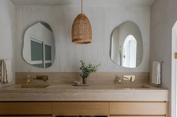 A refined contemporary bathroom vanity with a long double basin and timber drawer cabinetry beneath a beige stone benchtop and matching half-height wall finish. The upper wall features finely fluted vertical detailing, two organically shaped mirrors, and a woven pendant light. Brass-toned tapware and fittings add subtle warmth, while arched openings and reflected glazing contribute to the elegant character.