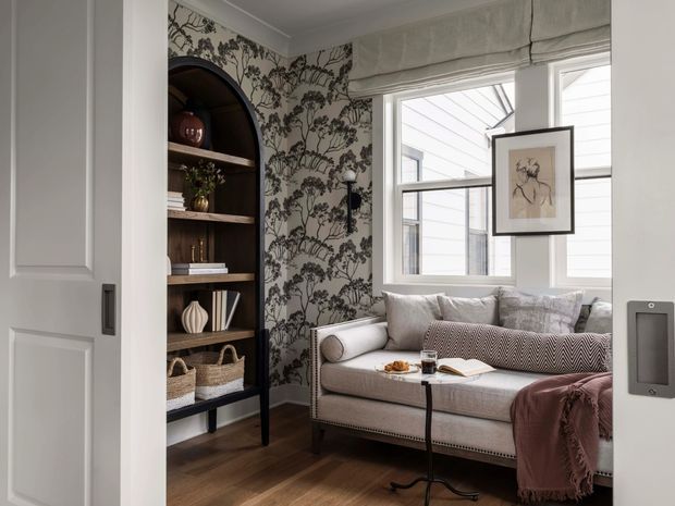 A refined, compact sitting room with a upholstered sofa positioned beside a large window, layered cushions, a throw blanket, and a small side table. A dark arched built-in shelving unit displays books and decorative objects, while botanical patterned wallpaper creates a dramatic feature wall. The room has warm timber flooring, white trim, soft Roman blinds, and understated traditional detailing.