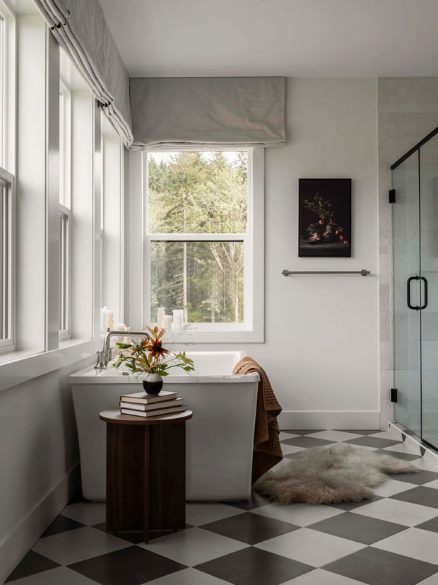 Bright, refined bathroom with a freestanding white bathtub positioned beneath a large window, a glass-enclosed shower, and a dramatic black-and-white checkerboard floor. Soft Roman blinds, light walls, a small dark wood side table, artwork, and a textured rug add warmth and character. The room overlooks a wooded setting.