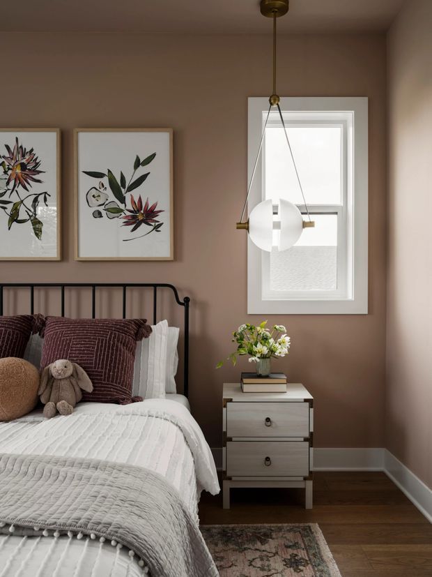 A warmly styled bedroom corner with a black metal spindle bed, layered white and grey bedding, muted burgundy pillows, framed botanical artwork, and a compact two-drawer bedside table. A sculptural white pendant light hangs beside a white-framed window. The room has taupe painted walls, dark timber flooring, and a small patterned rug, creating a calm, refined atmosphere.
