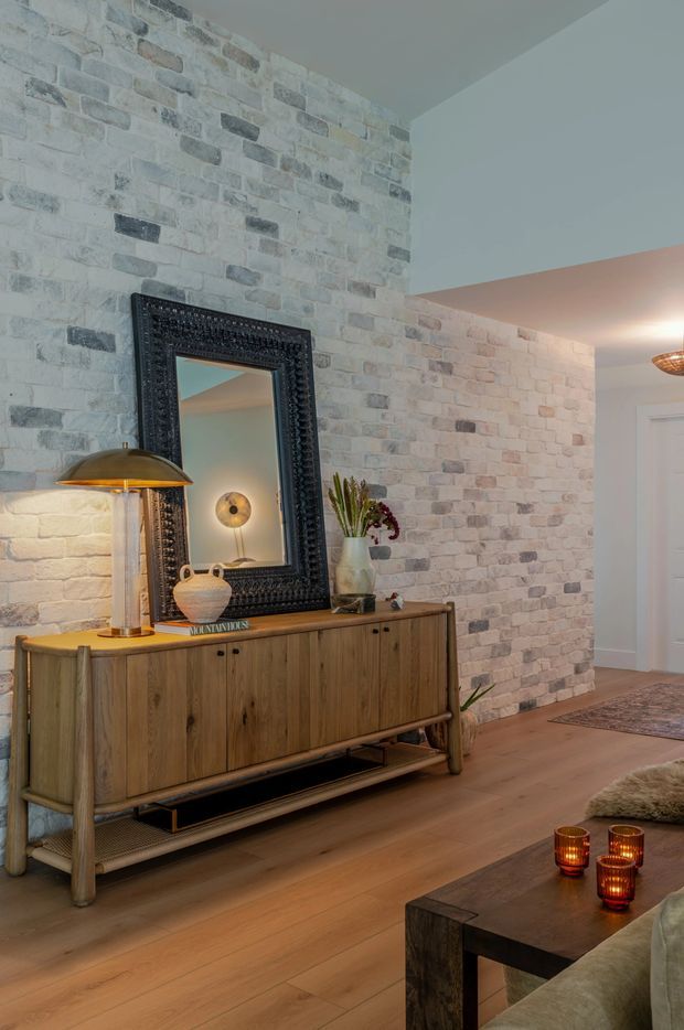 Warmly lit residential interior featuring an entryway or living-room transition with a whitewashed exposed brick feature wall, timber flooring, and a mid-century-style wooden sideboard with metal legs. A large ornate black-framed mirror, table lamp, decorative vase, plants, candles, and a partial sofa create a refined, inviting atmosphere. A hallway and doorway are visible beyond the feature wall.