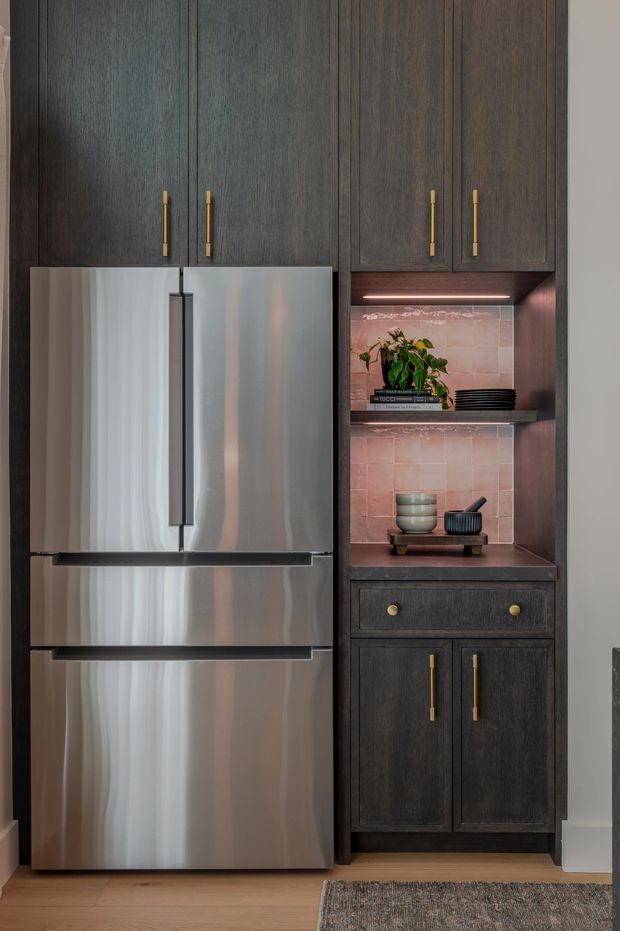 Close-up interior view of a compact kitchen storage wall featuring dark stained timber cabinetry with slim brass-gold pulls, a large stainless-steel refrigerator, and an illuminated open display niche. The niche has a pink-toned tiled backsplash, dark shelf, potted plant, stacked plates, and ceramic vessels. Light timber flooring and a neutral wall complete the refined, warm-toned composition.