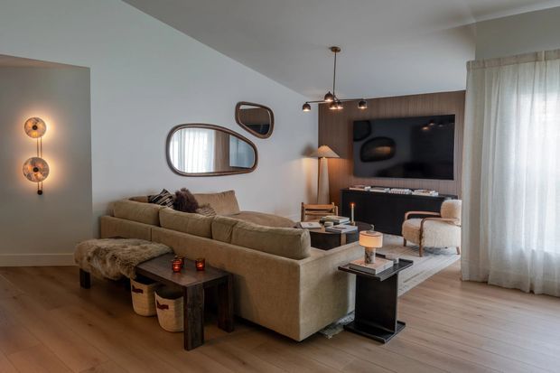 A warm, refined living room with a large beige sectional sofa arranged around a dark coffee table and side tables. The room features light timber flooring, white walls, sheer floor-to-ceiling curtains, a dark timber TV feature wall with built-in cabinetry, sculptural wall mirrors, layered pendant and wall lighting, and textured neutral furnishings.