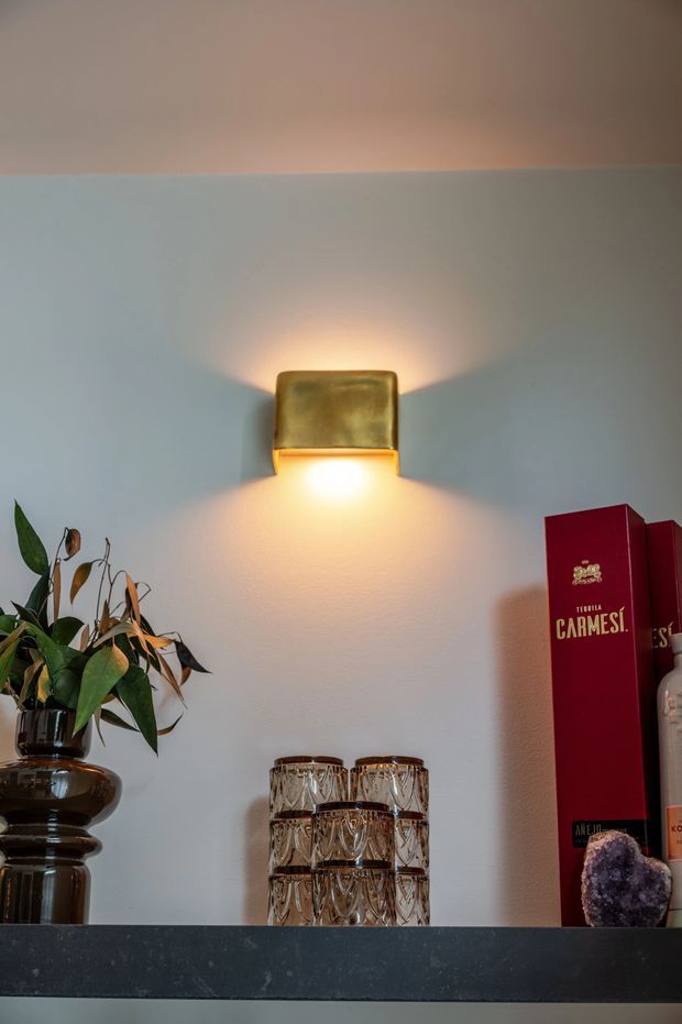 Close-up interior detail of a softly lit wall featuring a small brass-toned rectangular wall sconce casting warm light upward and downward. A dark stone or stone-look shelf runs across the lower portion, styled with a leafy plant in a dark sculptural vessel, stacked cut-glass tumblers, red books, and a decorative crystal. The scene has a refined, moody, warm atmosphere with minimal architectural detailing visible.