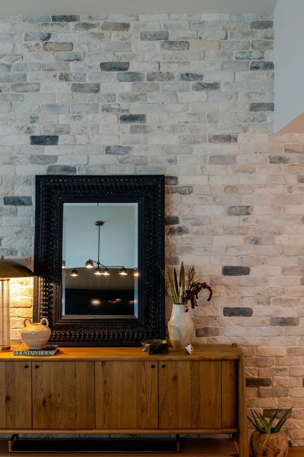 Interior vignette featuring a warm timber sideboard against a whitewashed exposed brick wall. A large ornate black-framed mirror reflects pendant lighting and a dark ceiling, while a ceramic vase, books, lamp, and plants add decorative detail. The composition has a refined, moody character with layered natural and dark finishes.