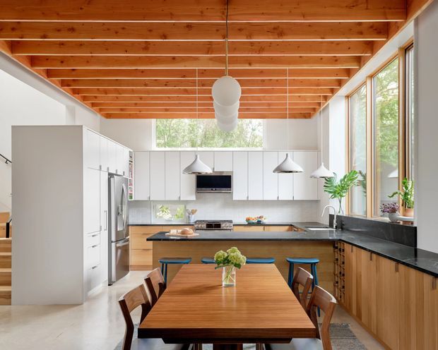 Bright open-plan kitchen and dining area with a warm exposed timber-beam ceiling, white full-height cabinetry, black stone benchtops, timber lower cabinetry, stainless-steel appliances, and a long timber dining table. Large windows provide abundant natural light and views to greenery, while pendant lights hang over the island and dining area.