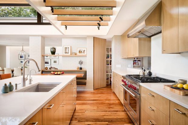 Bright open-plan kitchen with extensive light timber cabinetry, a large white island with integrated sink, stainless steel appliances, and warm timber flooring. The space opens to a living or dining area and features a glazed overhead roof opening with exposed timber beams and track lighting.
