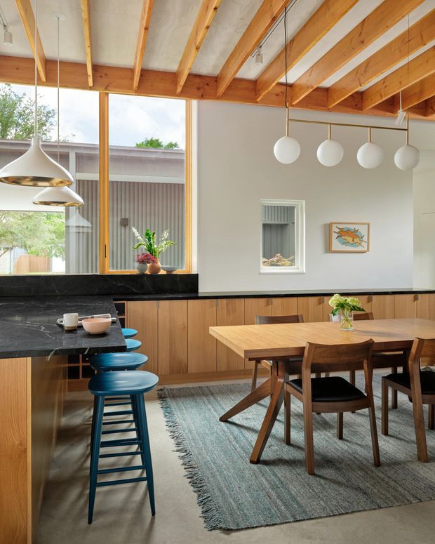 A bright, open-plan kitchen and dining area with extensive warm timber cabinetry, a black stone benchtop, exposed timber ceiling beams, and large timber-framed windows. A timber dining table with mid-century style chairs sits on a textured grey rug, while blue bar stools line the kitchen island. Sculptural pendant lights hang above the island and dining area. The room opens visually to an outdoor courtyard or garden beyond the glazing.