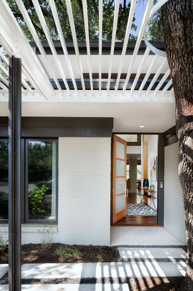 Exterior view of a contemporary home entry courtyard, featuring a white brick facade, dark-framed glazing, a timber-framed glazed front door, and a prominent white pergola with repeated overhead slats. A mature tree frames the right side, while the open doorway reveals a timber-floored interior entry with artwork and a patterned rug.