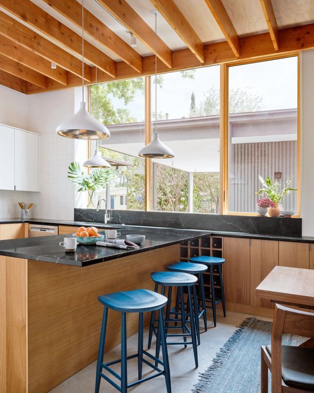 Bright contemporary kitchen with a large timber island and integrated seating, dark stone benchtops and splashback, white cabinetry, and a dramatic exposed timber-beam ceiling. Floor-to-ceiling glazing opens views to a covered outdoor area and garden beyond. Blue bar stools, pendant lights, plants, and a dining table complete the warm, airy interior.