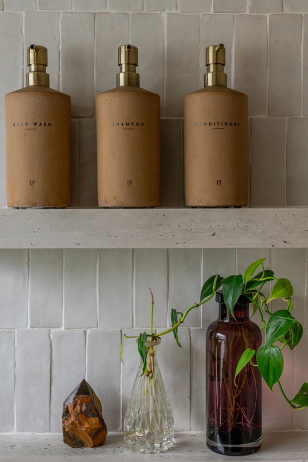 Close-up interior detail of built-in open shelving against pale handmade-look rectangular wall tiles. The upper shelf holds three matching amber-brown pump bottles labelled body wash, shampoo and conditioner, while the lower shelf displays a faceted stone, a clear glass propagation vase with plant cuttings, and a dark amber glass bottle containing a trailing green plant. The scene has a warm, earthy, curated bathroom or ensuite styling aesthetic.