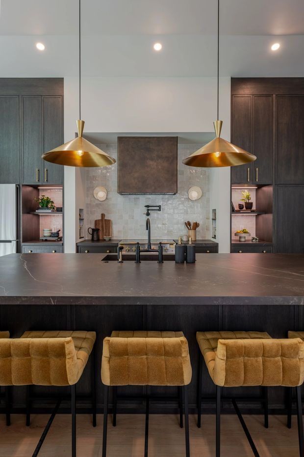A sophisticated contemporary kitchen viewed from the island side, featuring a large dark stone island with integrated sink and black tapware, dark timber cabinetry, a pale textured tile backsplash, and three mustard-brown upholstered bar stools. Two oversized brass pendant lights hang above the island, adding a warm metallic accent.