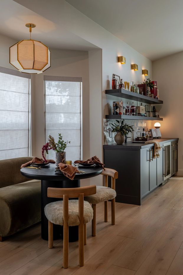 A refined dining and home bar area in a warm contemporary interior. A round black dining table with upholstered wooden chairs and a built-in bench sits beneath a large geometric pendant light. Behind it, dark built-in cabinetry with brass hardware supports open shelves displaying glassware and bottles, with brass wall sconces above. Light timber flooring, soft neutral walls, linen roller blinds, and layered warm lighting create an intimate entertaining space.