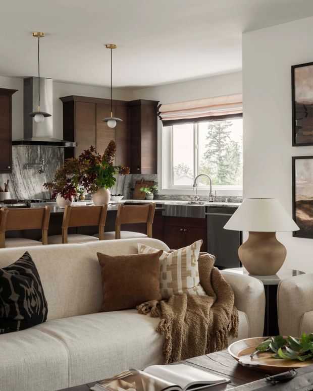 A warm, refined open-plan living and kitchen interior. In the foreground is a light neutral upholstered sofa with layered brown and patterned cushions and a textured throw, beside a sculptural ceramic table lamp. Behind it, a dark timber kitchen features marble-look stone surfaces, a substantial island with leather bar stools, pendant lighting, a sink beneath a large window, and botanical floral arrangements. Framed artwork and soft Roman blinds complete the inviting space.