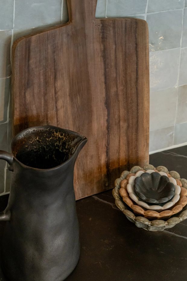 Close-up still life of a dark stone or concrete kitchen benchtop with a large warm-toned timber cutting board leaning against a pale grey tiled splashback. A matte black ceramic jug and a stack of textured decorative bowls sit in the foreground, creating a dark, earthy and tactile composition.