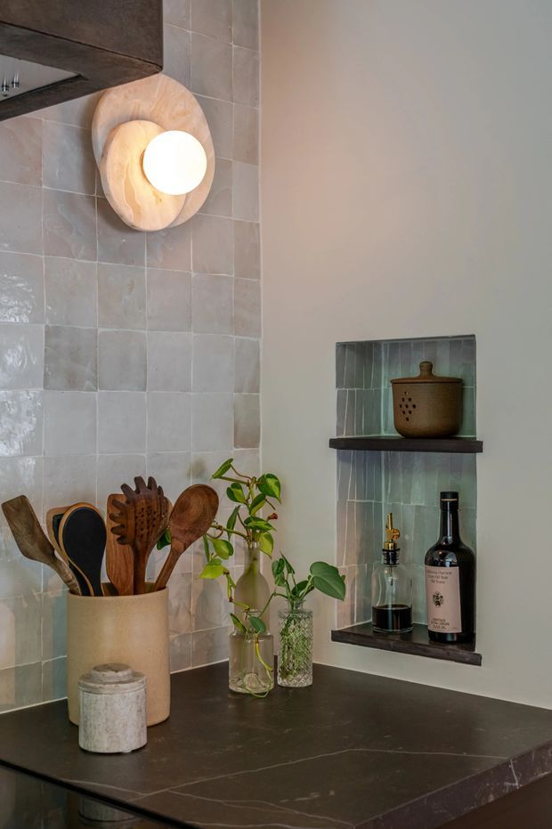 Close-up view of a warmly styled kitchen corner with a dark stone benchtop, pale square tiled splashback, integrated wall niches with dark shelves, timber cooking utensils, ceramic storage, greenery, and a sculptural wall light.
