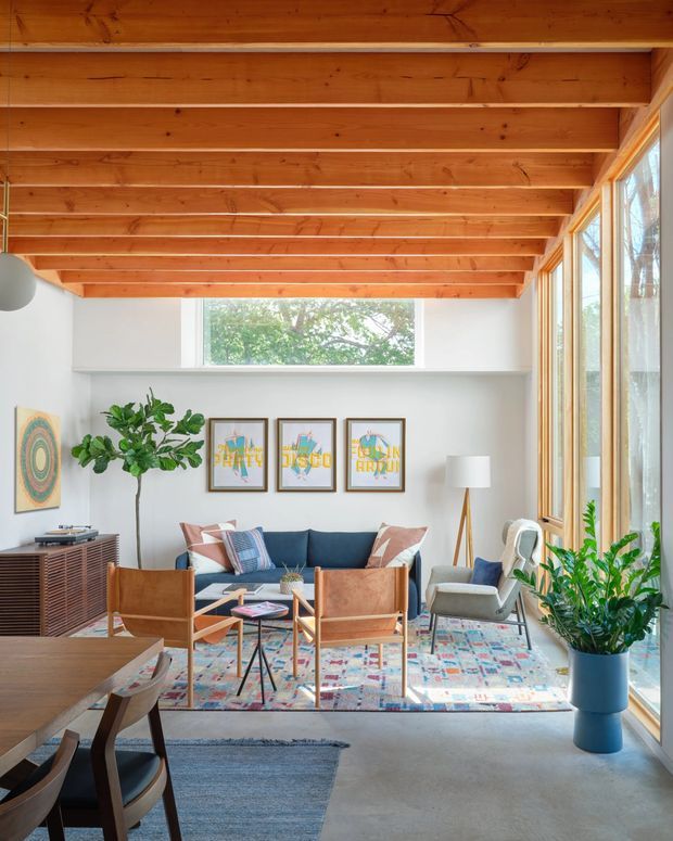 Bright, airy mid-century-inspired living room with exposed warm timber ceiling beams, white walls, expansive glazing, and a compact dining area in the foreground. The room contains a navy sofa, leather lounge chairs, patterned area rug, timber sideboard, framed artwork, indoor plants, and a tripod floor lamp. A large window and glazed wall provide views to greenery outside.