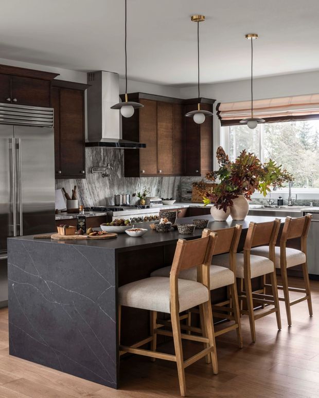 A sophisticated kitchen with a large dark stone island and integrated breakfast bar, surrounded by rich dark timber cabinetry. The island features subtle veining and is paired with several upholstered timber bar stools. A stainless-steel refrigerator, rangehood, cooktop, and marble-like stone splashback are visible, along with pendant lighting, a large window with a timber Roman blind, and a floral arrangement on the island. The palette is warm and refined, combining dark wood, charcoal stone, stainless steel, and light timber flooring.