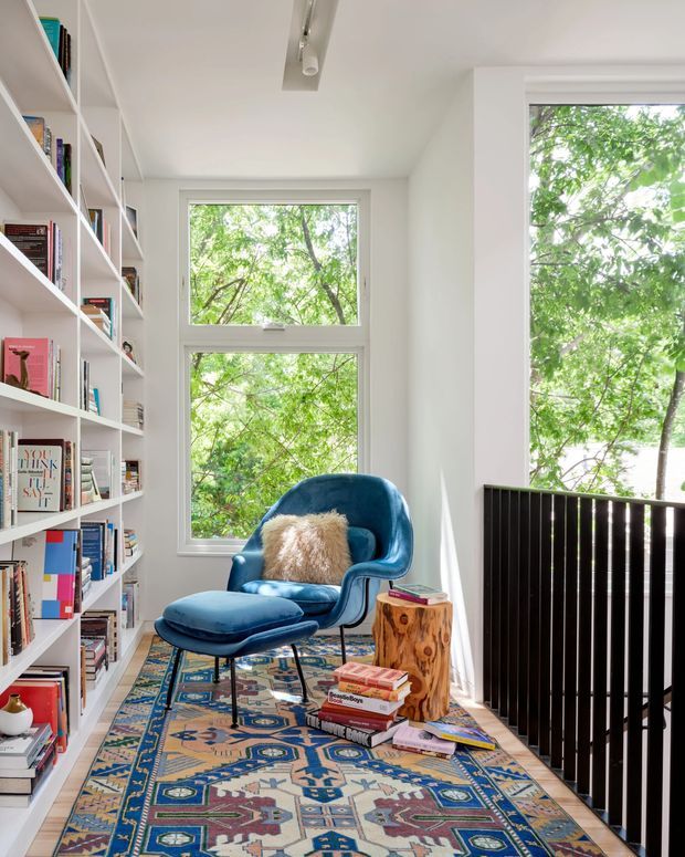 Bright, narrow reading nook or home library with floor-to-ceiling white built-in bookshelves, large windows overlooking leafy greenery, and a deep blue velvet lounge chair with matching ottoman. A colourful patterned rug, rustic sculptural wood side table, and stacks of books create a warm, eclectic retreat. Black vertical balustrading borders the right side, suggesting the space is on an upper-level landing or mezzanine.