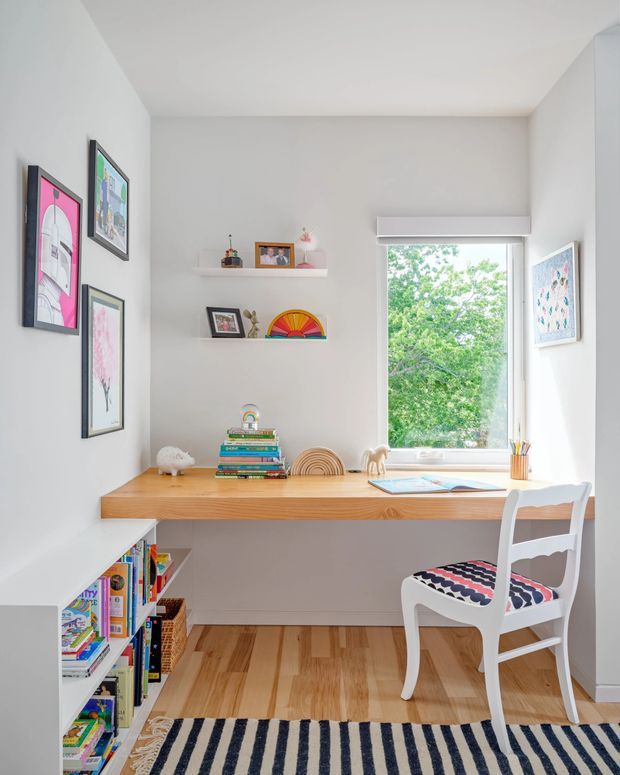 Bright, compact home office or study nook with a built-in timber desk spanning beneath a large window. White walls and shelving display colourful framed artwork, books, toys, and small decorative objects. A white wooden chair with a patterned upholstered seat sits at the desk, while a striped rug and warm timber flooring add contrast. The space feels playful, tidy, and inviting, with abundant natural light.