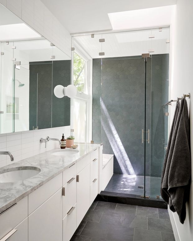 Bright contemporary bathroom with a long white vanity, pale marble benchtop, integrated basins, large wall mirror, glass-enclosed walk-in shower, dark grey hexagonal shower tiles, and dark grey floor tiles. A skylight and tall window provide abundant natural light, while brushed metal hardware and dark towels add contrast.
