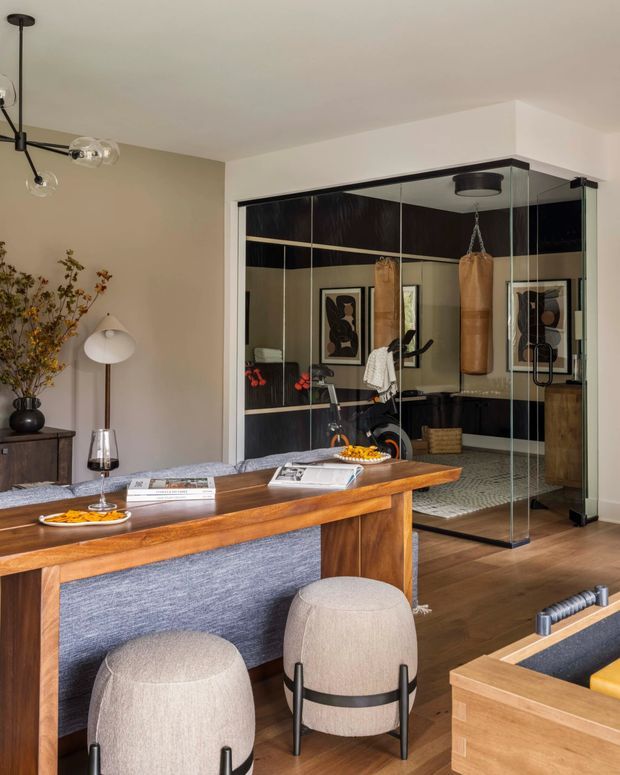 A refined open-plan living and dining area with warm timber flooring and a substantial timber dining table positioned behind a blue-grey upholstered sofa. Two rounded upholstered stools sit in the foreground beside a light timber cabinet. The room features beige walls, dark timber or black built-in cabinetry, framed artwork, contemporary lighting, and a fully glazed home gym enclosure with a hanging punching bag in the background.