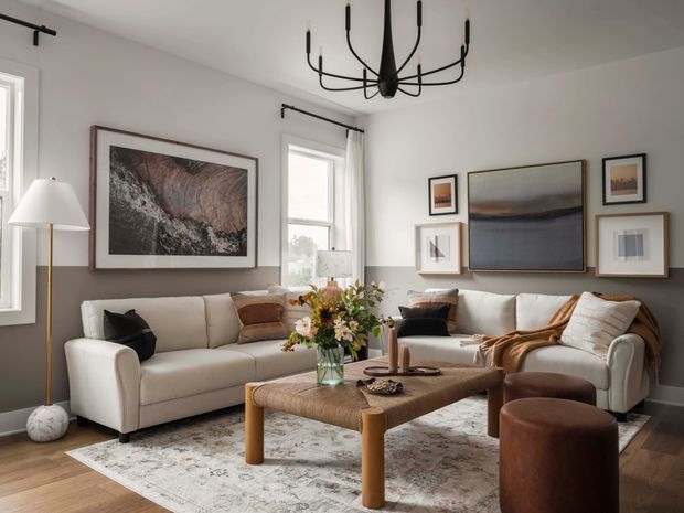 A refined living room with a neutral grey and cream palette, two upholstered sofas, a large textured rug, and a substantial woven or upholstered rectangular coffee table. The walls feature a curated arrangement of framed abstract artwork, while a sculptural black multi-arm chandelier provides a strong focal point. Timber flooring, white-trimmed windows, floor lamp, cushions, and floral styling complete the warm contemporary setting.