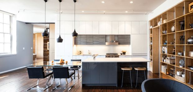 A bright, open-plan contemporary kitchen and dining area. The kitchen features a large dark island with a pale benchtop, white full-height cabinetry, a timber-toned tiled or panelled splashback, integrated appliances and three black pendant lights. A dining table with dark upholstered chairs sits beside the kitchen, while an extensive open timber display shelving unit lines the right wall. Large windows provide abundant natural light.