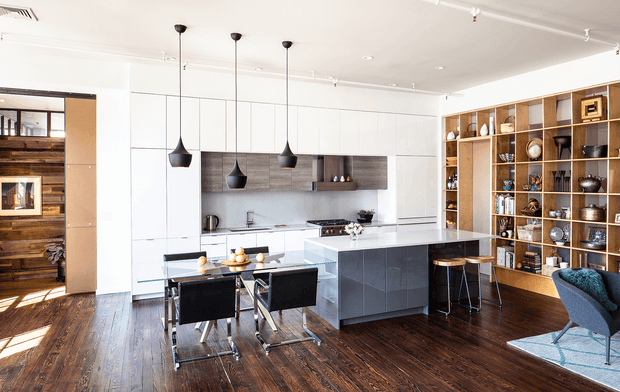 Bright open-plan kitchen and dining area with a large white island, dark integrated cabinetry, black pendant lights, timber flooring, a compact dining table, and a full-height timber display shelving wall. A wood-burning fireplace and adjoining room are visible through the opening at left.