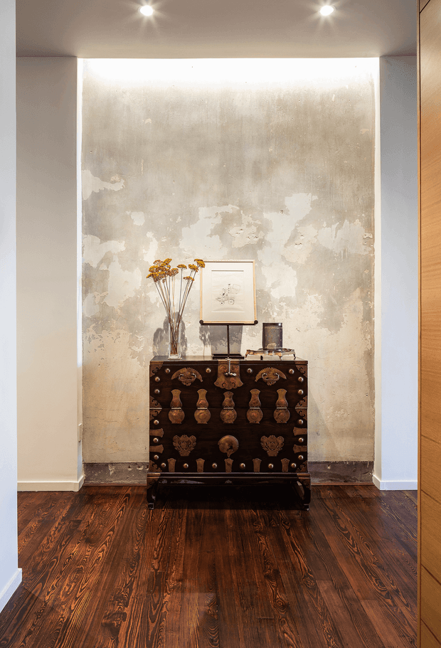 A styled interior alcove or hallway vignette featuring an ornate dark wood antique chest against a distressed textured plaster feature wall. The chest is decorated with metal hardware and topped with a tall glass vase of dried flowers, framed artwork, and small decorative objects. Dark timber flooring, recessed ceiling spotlights, white wall returns, and a warm timber panel or door frame the composition.