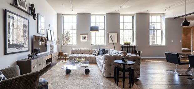 A spacious, light-filled open-plan living room with tall industrial-style multi-pane windows, grey walls, dark timber flooring, and a large tufted sectional sofa arranged around a low coffee table. Framed artwork, sculptural decor, side tables, and a compact dining area are visible, creating a refined contemporary loft-like interior.