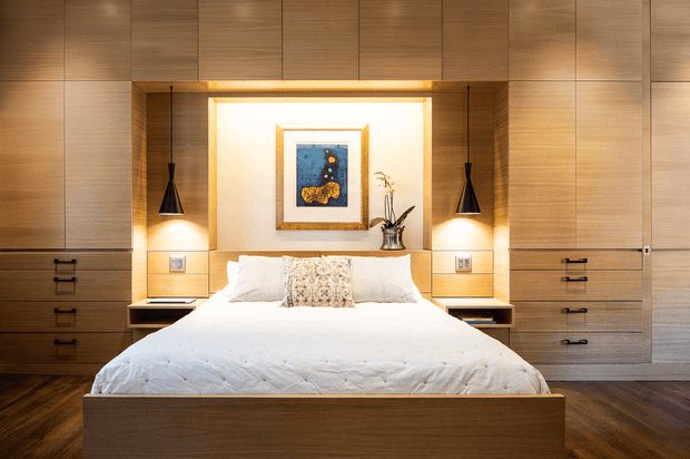 A warm, symmetrical contemporary bedroom featuring a built-in timber bed surrounded by full-height timber cabinetry. Integrated bedside niches, black pendant lights, a softly illuminated recessed art display, and dark timber flooring create a refined, intimate atmosphere.