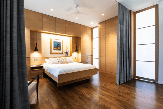 A warm, contemporary bedroom with a floating timber bed, extensive built-in timber cabinetry and wall panelling, integrated bedside storage, and illuminated bedside niches. Dark timber flooring, tall frosted-glass windows or doors, and full-height charcoal curtains create a refined, softly lit interior. A ceiling fan, recessed lighting, artwork, and layered bedding complete the room.