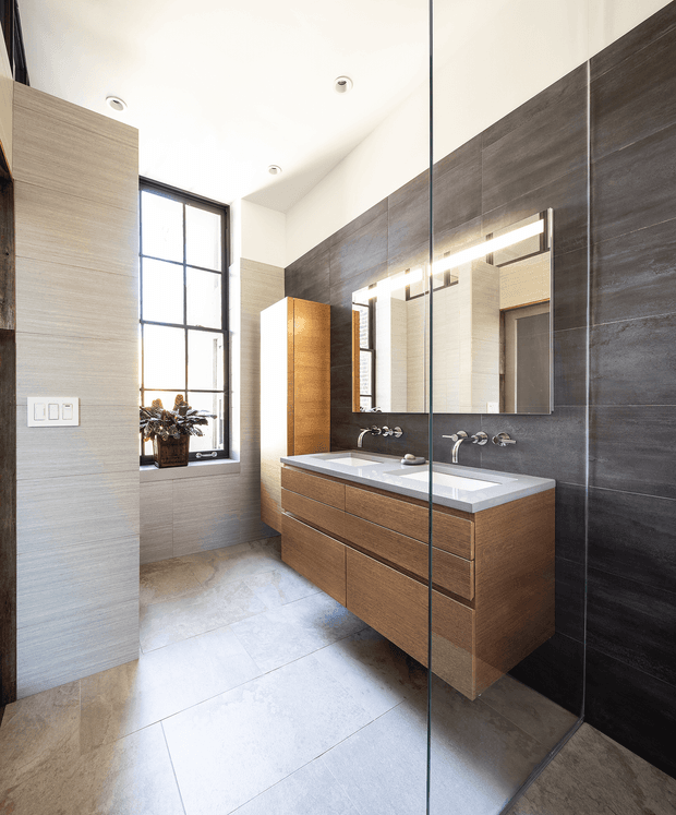 Contemporary bathroom featuring a long double vanity with integrated rectangular basins, timber-look drawer cabinetry, pale stone or solid-surface benchtop, large-format grey wall and floor tiles, and a full-height frameless glass shower screen. A large illuminated mirror spans the vanity, while a tall black-framed window brings in natural light.