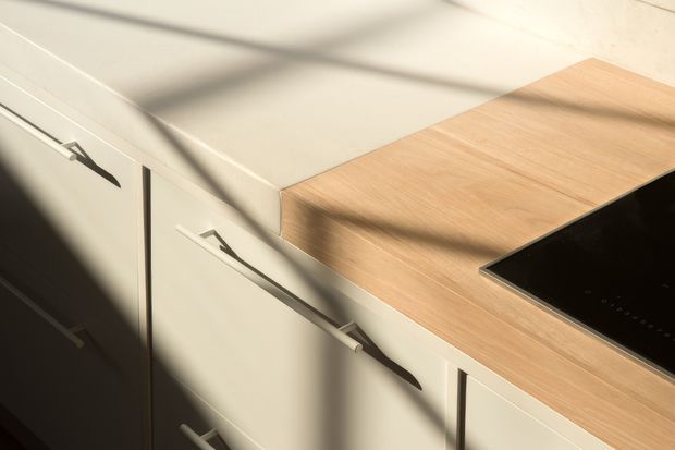 Close-up of a contemporary kitchen work area with pale cabinetry, slim dark metal handles, a light timber benchtop, and a black glass induction cooktop. Strong sunlight casts geometric shadows across the cabinetry and countertop.