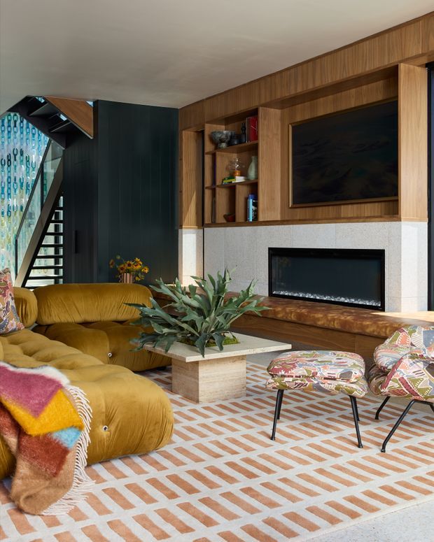 A richly layered contemporary living room with a mustard-gold curved sectional sofa, patterned occasional chairs, a geometric patterned rug, and a central stone-topped coffee table. The room features extensive warm timber built-in cabinetry and shelving around a dark fireplace and television, a pale stone or concrete fireplace surround, dark vertical wall panelling, and a dramatic open stair with patterned glazing. The scheme combines warm wood, ochre upholstery, colourful textiles, and bold geometric details.