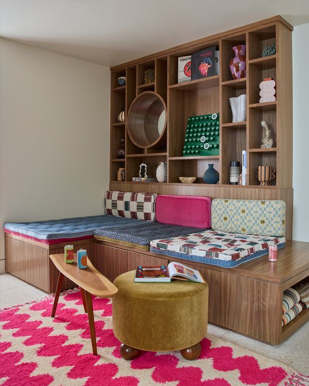 A colourful mid-century-inspired built-in banquette occupies the corner of a room, with upholstered patchwork seat cushions and backrests integrated into warm wood cabinetry. Above it is a large open timber display shelving unit filled with ceramics, books, artwork and decorative objects. A small curved wooden coffee table, mustard upholstered ottoman, and bold pink-and-cream patterned rug complete the inviting reading or lounging nook.