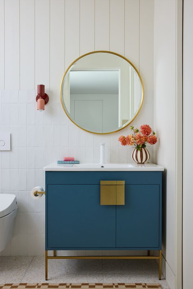 A stylish compact bathroom featuring a floating deep blue vanity with brass hardware and slender brass legs, a white countertop basin, white square tiled lower walls, and vertically panelled white walls above. A large round mirror with a gold frame hangs over the vanity, accompanied by a red and pink wall sconce. A patterned terrazzo-like floor, toilet, and floral arrangement complete the colourful contemporary scheme.