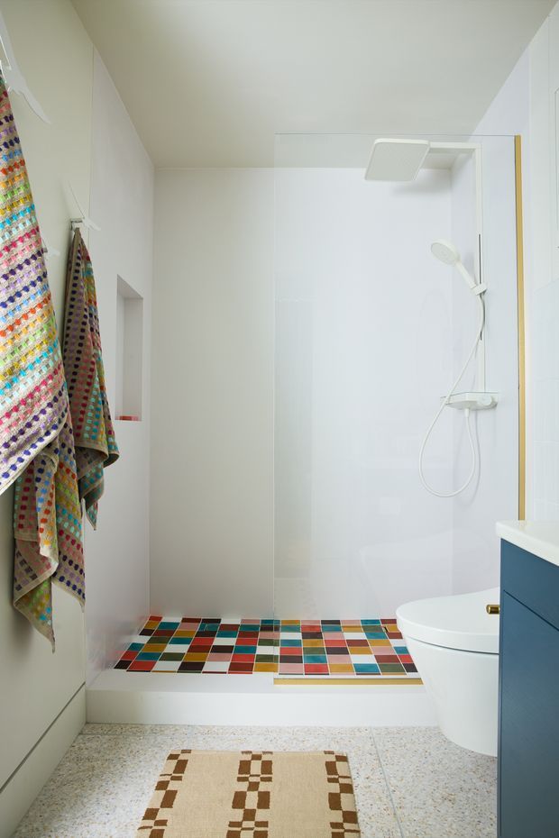 Bright compact bathroom with a white walk-in shower, wall-mounted shower fixtures, white toilet, and a highly colourful geometric mosaic tile floor in the shower area. A terrazzo-like light floor continues into the room, with colourful patterned towels on wall hooks, white cabinetry, and a dark blue vanity edge visible at right.