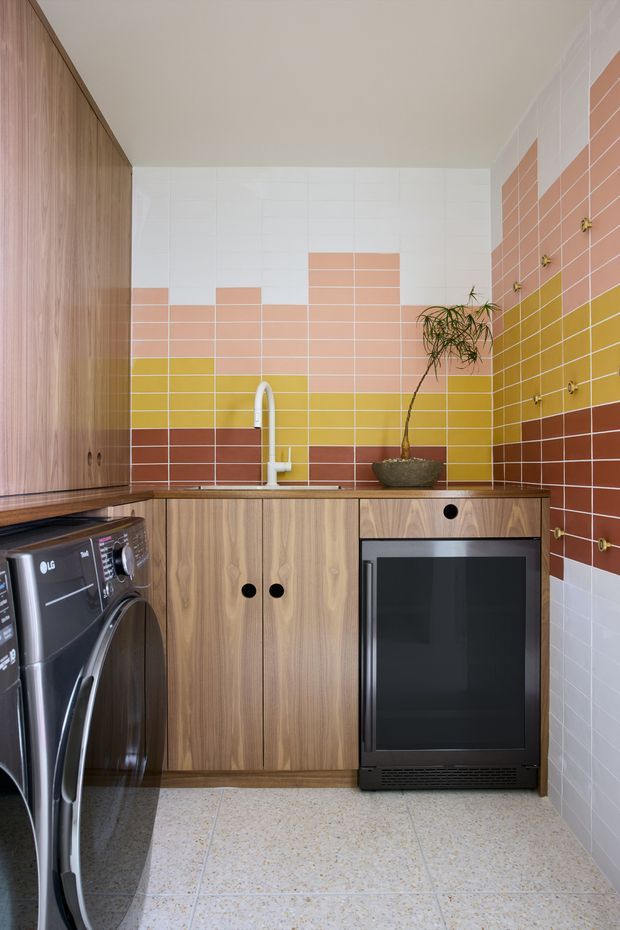 Compact, colourful laundry room with full-height timber-look cabinetry, a front-loading washing machine, integrated dark appliance, sink, and a potted plant. The walls feature a playful geometric arrangement of white, peach, mustard yellow, and terracotta rectangular tiles, while the floor is finished in pale terrazzo-style tiles.