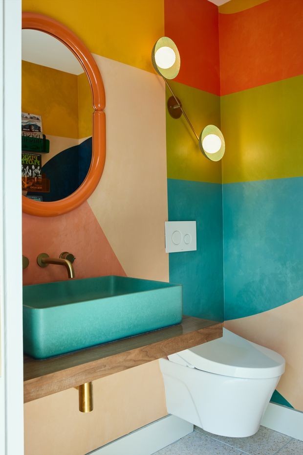 A compact, highly colourful bathroom with bold geometric painted wall sections in orange, mustard yellow, olive green, teal, peach, cream and navy. The room features a wall-mounted teal vessel basin on a timber vanity shelf, a brass wall-mounted tap and exposed brass waste, a large rounded orange-framed mirror, decorative wall sconces, a white wall-hung toilet, and terrazzo-style flooring. The design is playful, graphic and eclectic with strong retro influences.