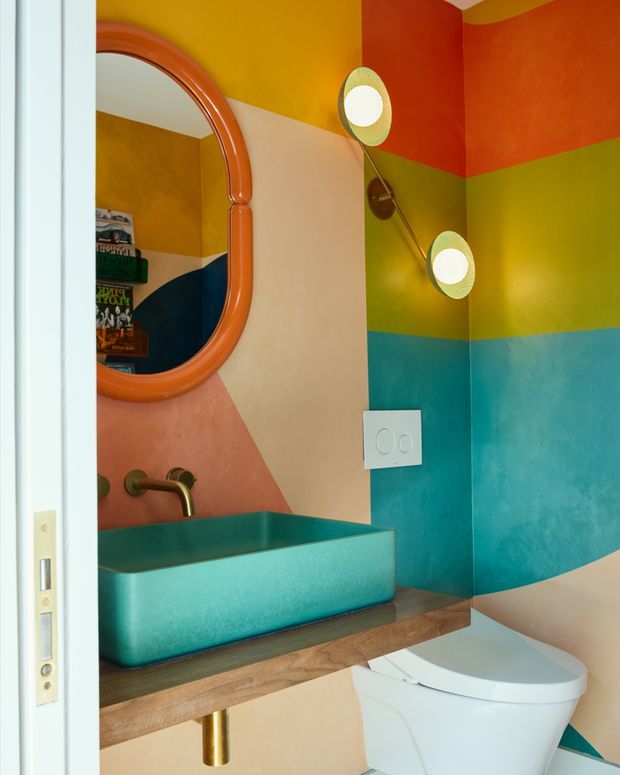 A compact, vividly coloured bathroom viewed through an open doorway. The room features a turquoise vessel basin on a wall-mounted timber vanity shelf, a white wall-hung toilet, brass tapware and drain, a rounded orange-framed mirror, and twin globe wall lights. The walls are decorated with bold horizontal and geometric bands in orange, mustard yellow, olive green, turquoise, peach, and cream.