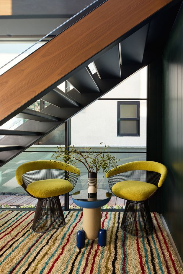 A dramatic contemporary interior showing a sculptural black metal stair with a warm timber-clad underside and clear glass balustrade. Two curved mustard-yellow lounge chairs with black wire bases flank a small round table, set on a colourful striped rug. Glazing, a planted courtyard view, and dark vertical wall panelling add texture and contrast.