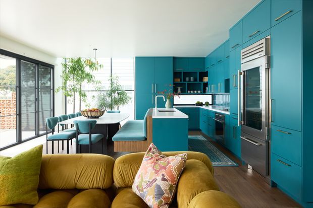 Bright open-plan kitchen and dining area connected to a living space. The kitchen features extensive turquoise-blue fixed cabinetry, integrated stainless-steel appliances, a white island benchtop, open shelving, and a patterned backsplash. A compact dining table sits beside large black-framed glazed doors, while the foreground contains a mustard-yellow sofa with a colourful patterned cushion. Timber flooring and abundant daylight create a vibrant, contemporary interior.