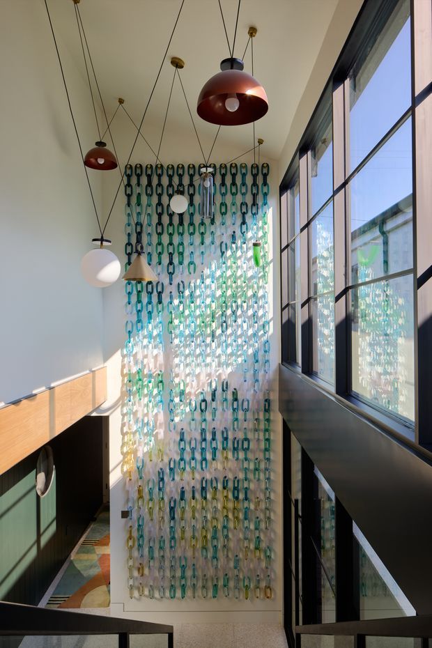 A tall, light-filled interior stairwell or landing with a dramatic double-height void. Large black-framed windows line the right side, while a full-height decorative screen made of translucent turquoise, green, and amber chain links forms a striking feature wall. Several suspended globe and dome pendant lights hang at different heights, and dark metal stair or balustrade elements frame the lower portion of the image. Warm sunlight creates strong reflections and shadows across the pale walls and floor.