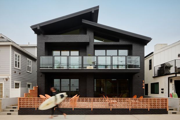 Street-facing view of a contemporary multi-storey residence with a dramatic asymmetrical pitched roof, dark brick facade, broad glazing, and a glass-railed upper balcony. A distinctive terracotta breeze-block screen encloses the front terrace, with neighboring urban homes closely flanking the property.